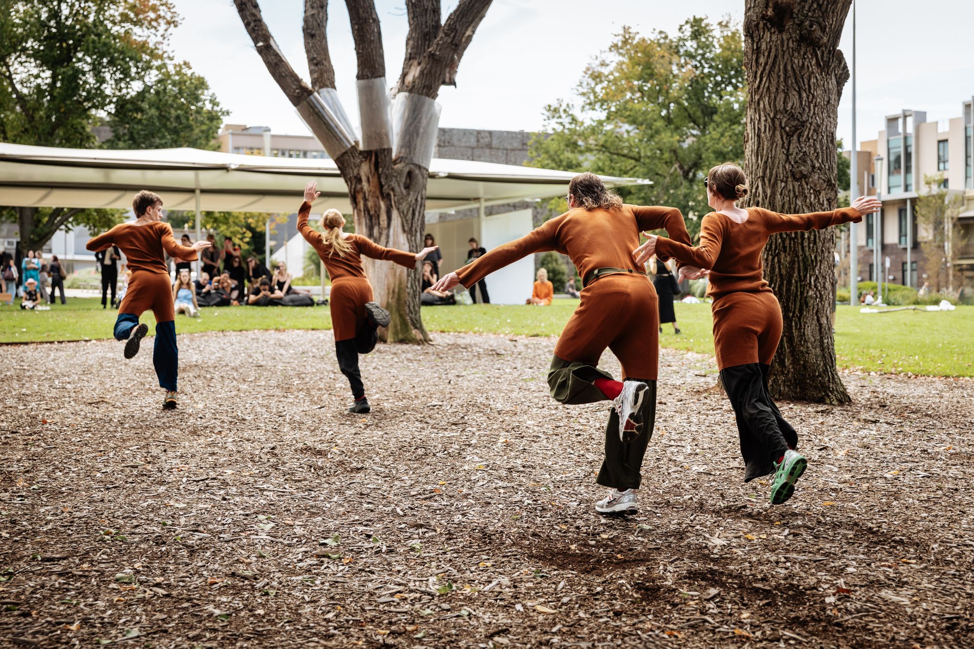 A photo of four performers in brown overalls are moving away from the camera towards an outdoor pavilion in the park