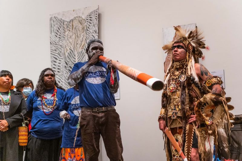 A Yolnu man plays a didgeridoo in a gallery. A native american indian man listens next to him.