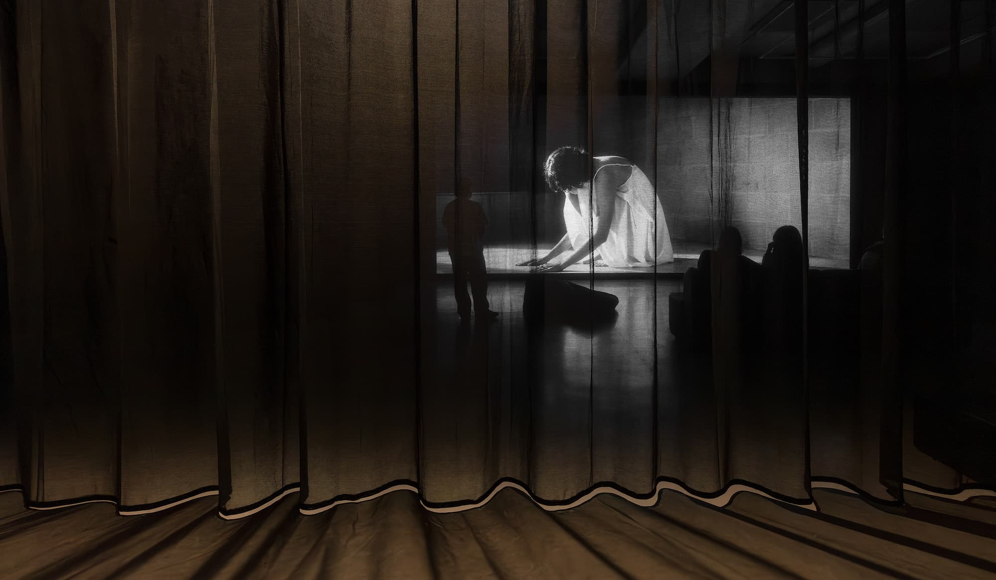 A person in a white dress crouches down on a large black and white screen with a translucent curtain in foreground