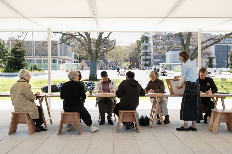 A group of people sit talking at a table and doing an activity in an open space building in a garden