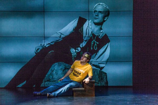 An actor on stage lazes on a chaise lounge with a projection behind