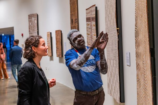 A Yolnu man with traditional face paint speaks to a visitor about an artwork 