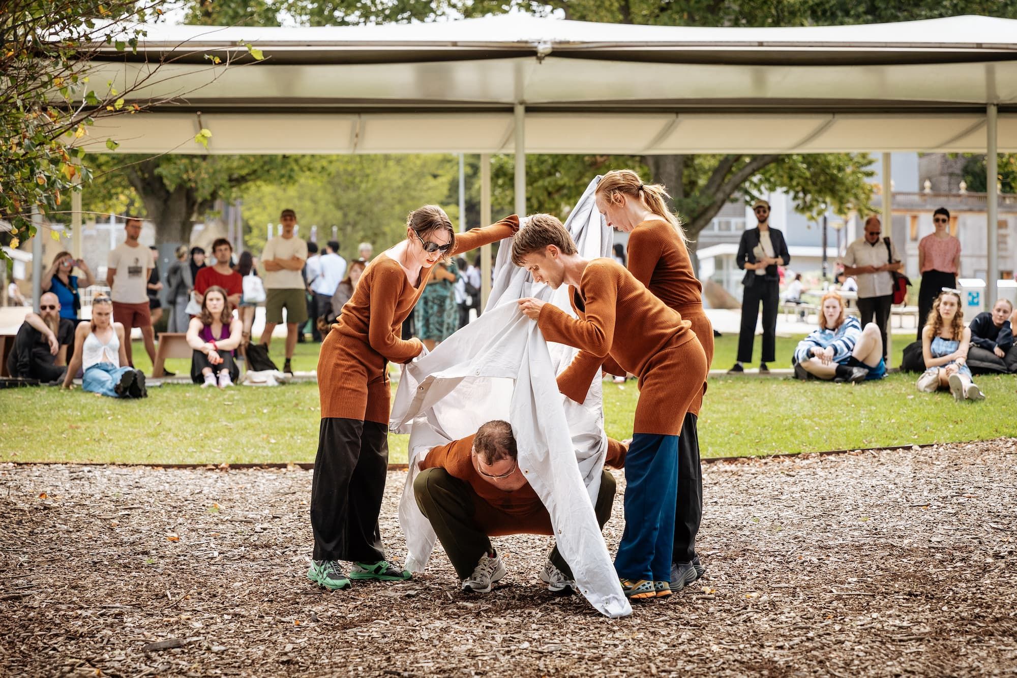 A group of performers are lifting up a white piece of fabric in front of an outdoor pavilion