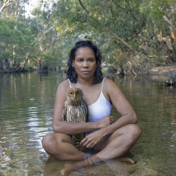 A woman sits in a river with an owl resting on her arm