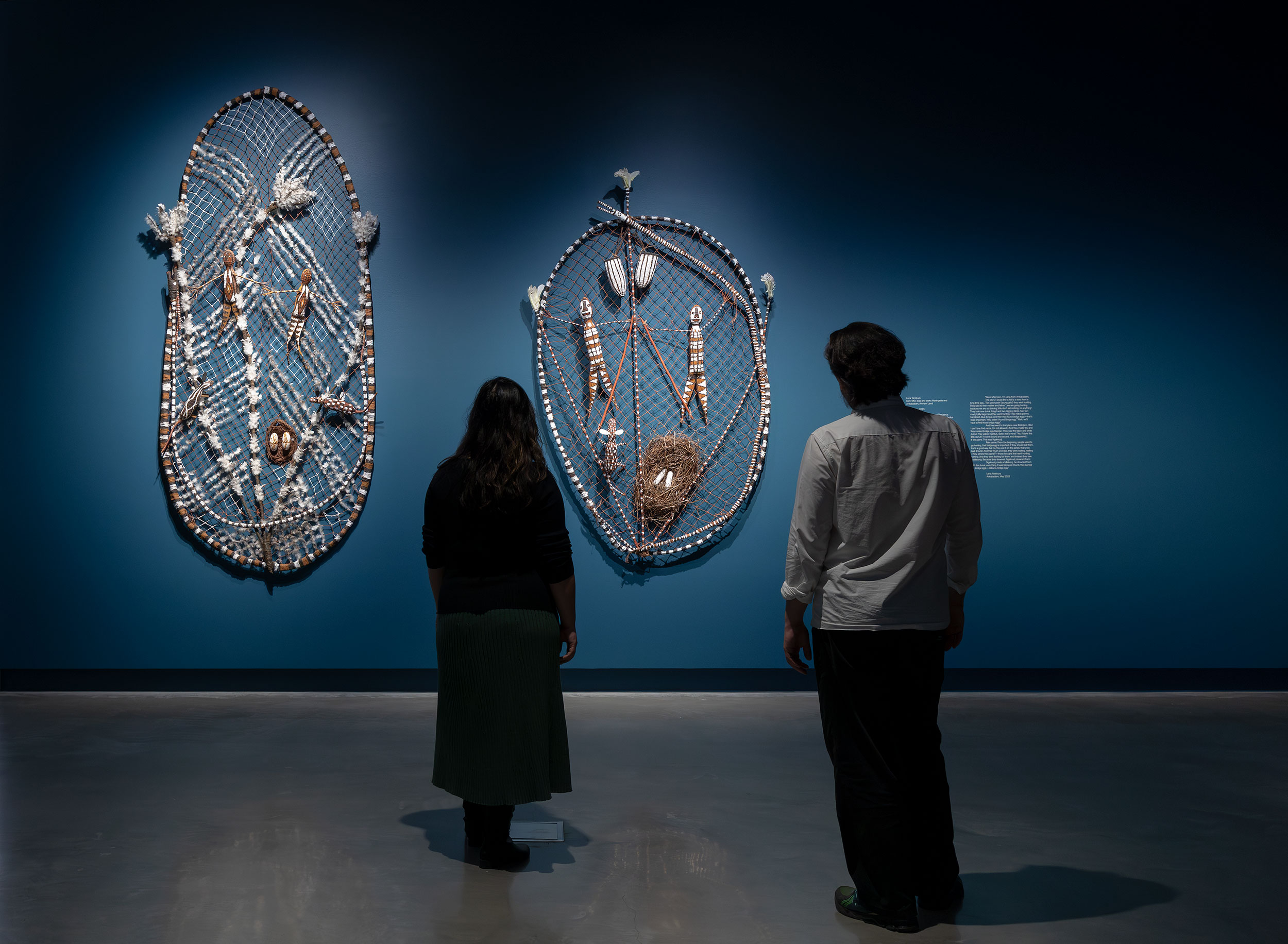 Two people look at a feathered round sculpture on a blue wall