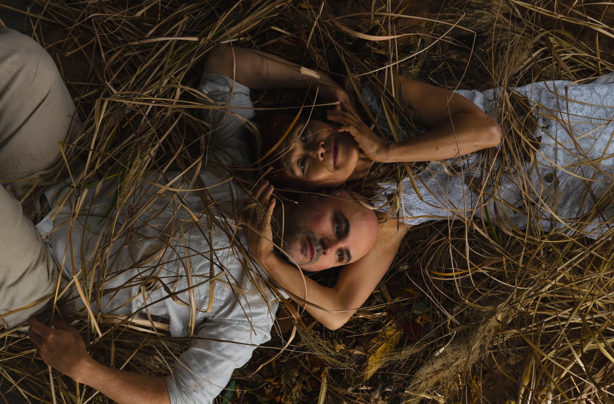 Two people lie on a bed of straw head to head arms wrapped around each other