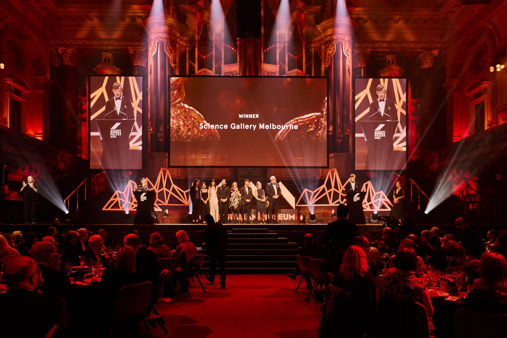 A group of people in formal attire stand arm in arm on a stage in a large auditorium