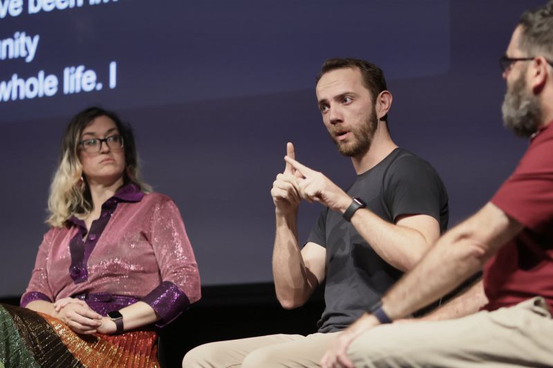 Three people sit on a stage and person in middle signs
