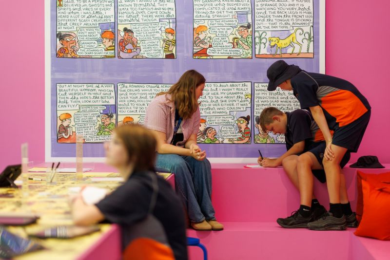 Two students write in a book as a teacher looks on