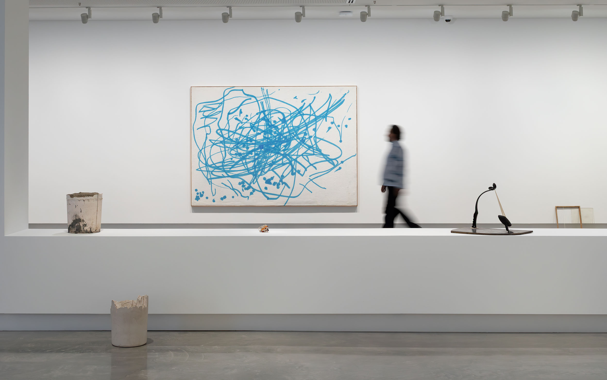 A person walk in front of a blue scribble in a gallery with small sculptures in foreground