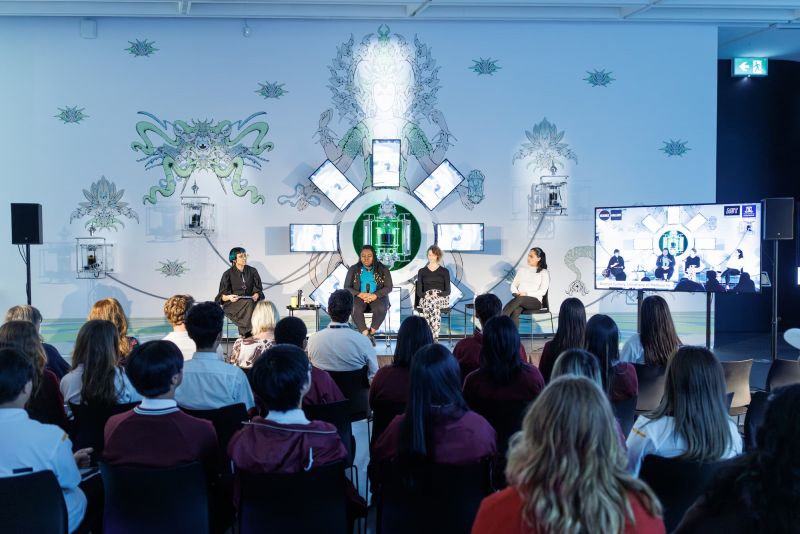 People watch presenters on a stage in a gallery