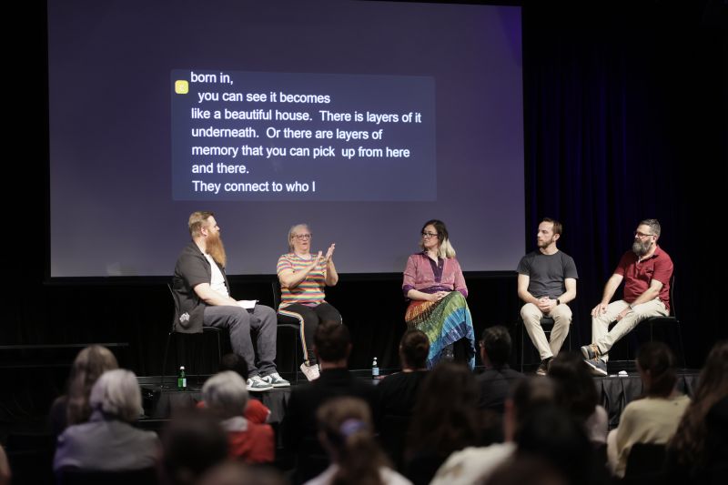 A number of people sit on a stage in front of an audience. One person is signing