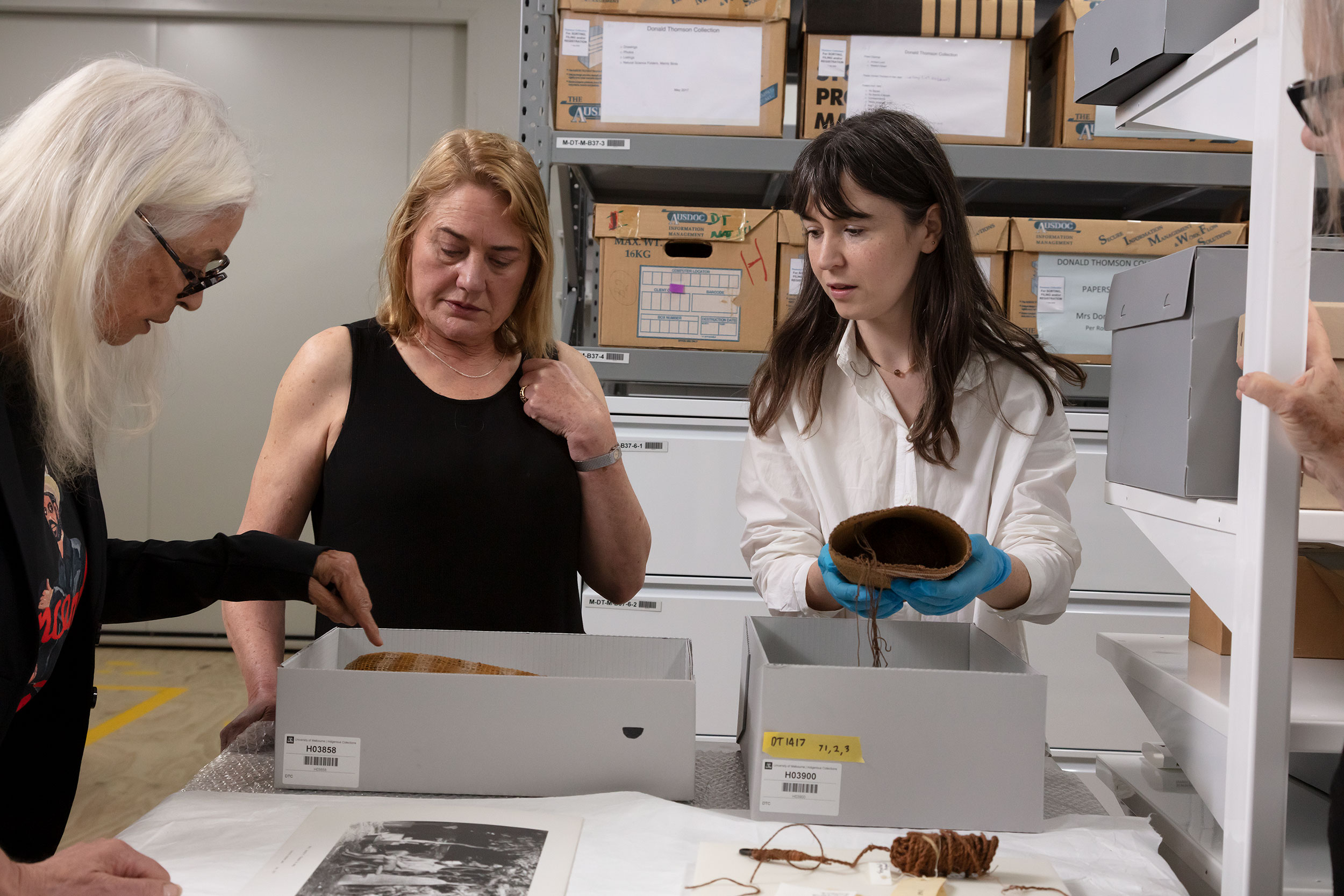 Three people are looking at a woven basket located in a protective box on the table in the collection store. The person on the right holding another basket in their gloved hands