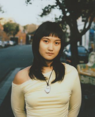 a person with black hair and a fringe and shoulder length hair wears a yellow off the shoulder top with a shell neklace. street soft in background.