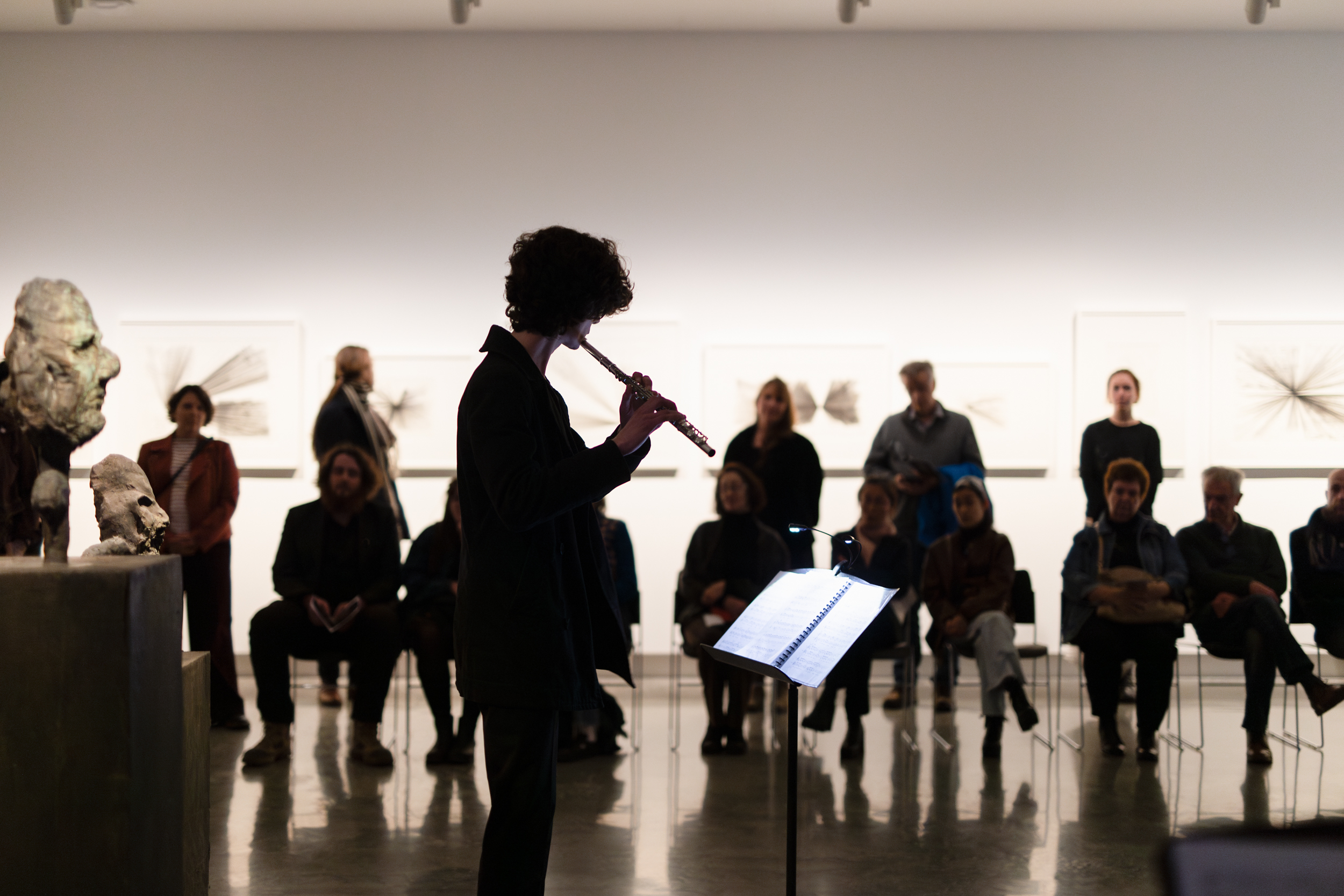 A person plays a flute in front of an audience in a gallery